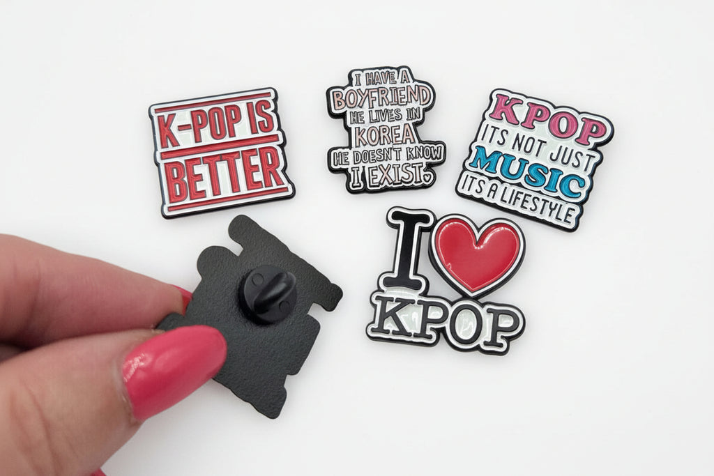 Four K-pop themed pins held by a hand with pink nail polish on a white background.